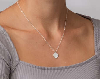 Puede incluir: Un collar de plata con un pequeño colgante circular suspendido de una cadena delicada. El collar tiene una longitud total de cadena de 45,72 cm. El metal tiene un acabado pulido y el dije circular cuelga centrado en la base del cuello.