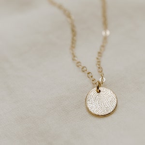 Puede incluir: Un collar de oro con un colgante redondo que presenta un dise&ntilde;o detallado de huella digital. El colgante tiene un di&aacute;metro de aproximadamente 2,5 cm. El collar est&aacute; sobre un fondo de tela blanca. La imagen tambi&eacute;n muestra un primer plano del dise&ntilde;o de la huella digital, etiquetado como "huella digital real".