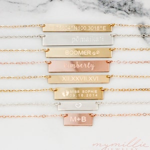 May include: A set of eight silver, gold, and rose gold bar necklaces with various engravings. The engravings include coordinates, names, dates, symbols, and Roman numerals. The necklaces are on a white marble background.