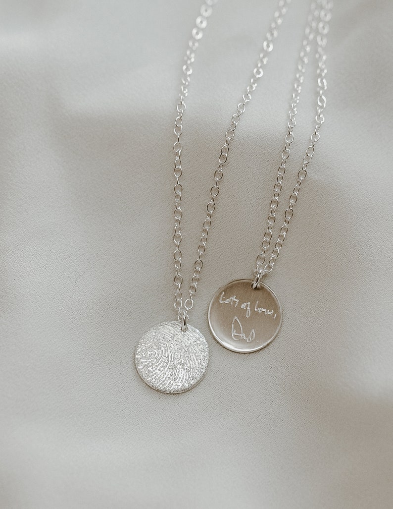 May include: Two silver necklaces with circular pendants. One pendant features a fingerprint design, the other has the handwritten message "Lots of love, Dad".