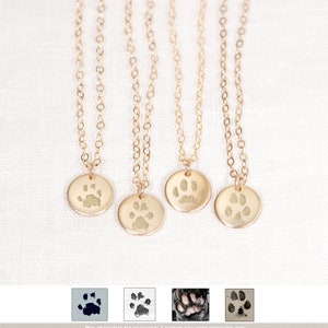 May include: Four gold necklaces with a small round pendant featuring a paw print design. The pendants are engraved with a custom paw print. The necklaces are displayed on a white fabric background.  Max characters for engraving: 1 custom paw print engraving only. Photos of Real Paws, Inked Prints, & Clay Impressions accepted.