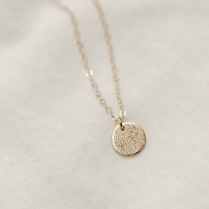 Puede incluir: Un collar de cadena de oro con un colgante redondo que presenta un dise&ntilde;o detallado de huella digital.