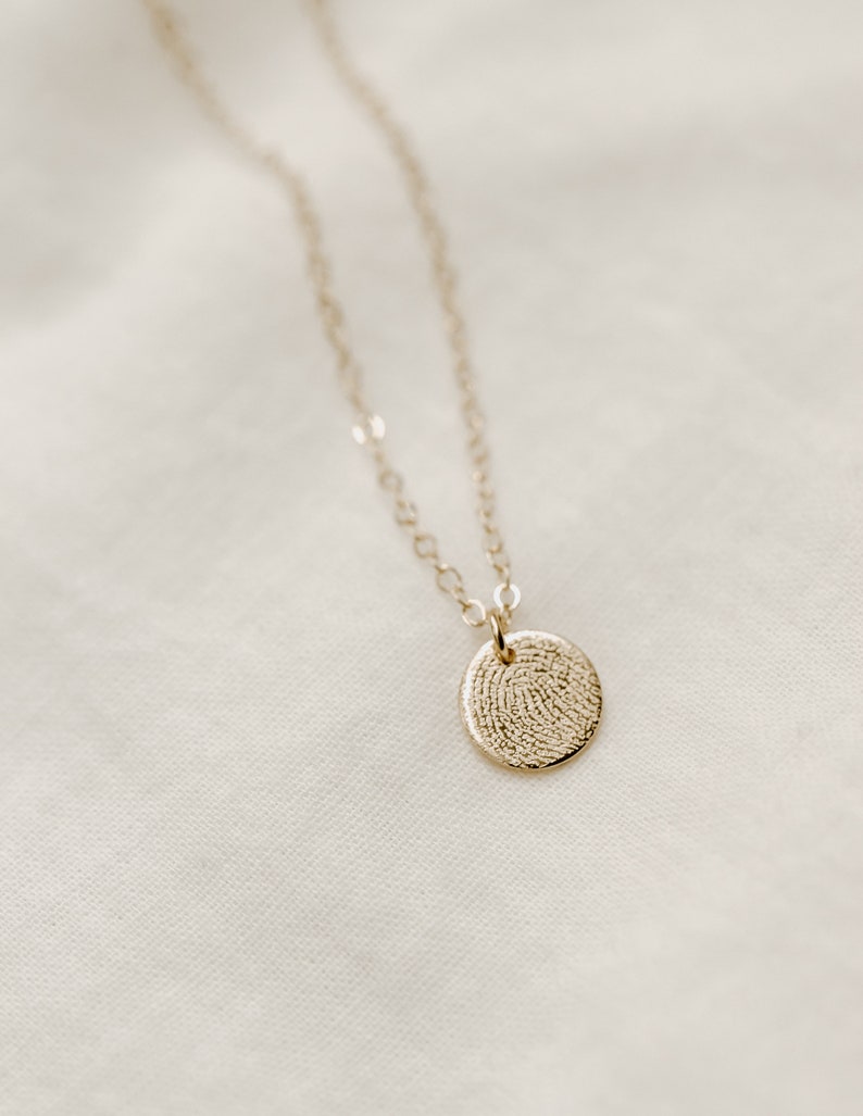 Puede incluir: Un collar de cadena de oro con un colgante redondo que presenta un dise&ntilde;o detallado de huella digital.