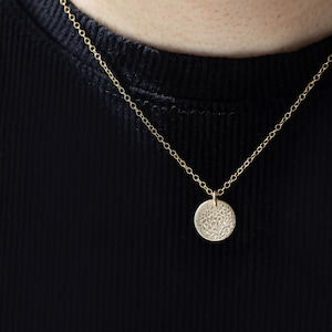 Pode incluir: Um colar dourado com um pingente circular texturizado. O pingente pende de uma corrente delicada. O fundo &eacute; um tecido preto canelado.
