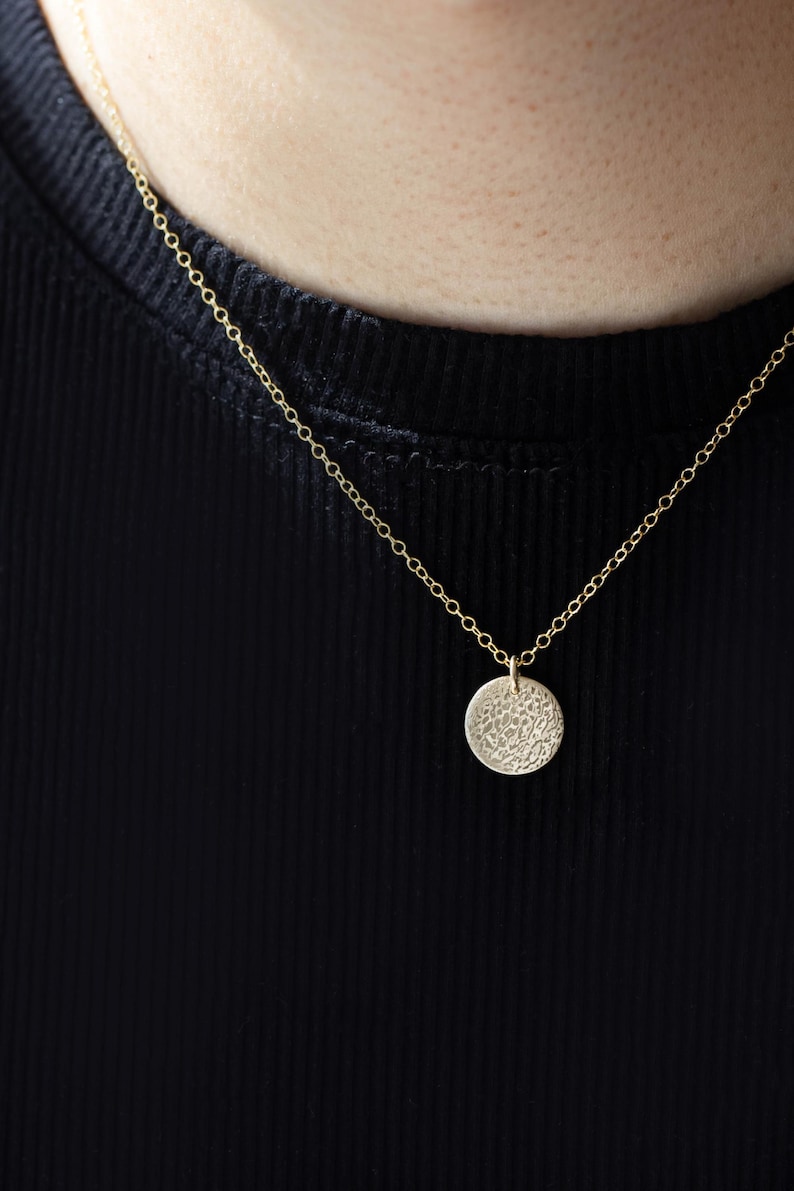 Pode incluir: Um colar dourado com um pingente circular texturizado. O pingente pende de uma corrente delicada. O fundo &eacute; um tecido preto canelado.