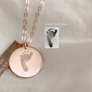 May include: A rose gold necklace with a small round pendant featuring a baby's footprint. The pendant is attached to a delicate chain. The necklace also includes a small rectangular tag with the date "07.14.18" engraved on it.