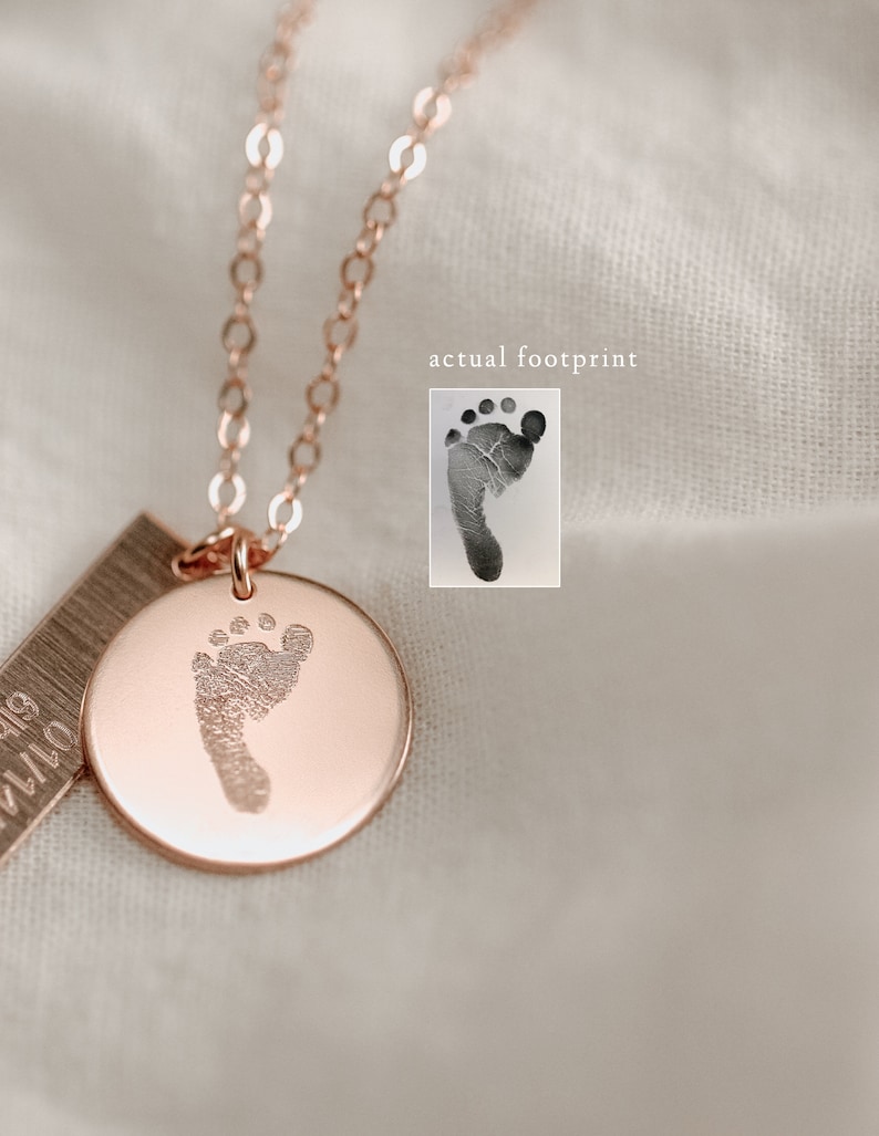 May include: A rose gold necklace with a small round pendant featuring a baby's footprint. The pendant is attached to a delicate chain. The necklace also includes a small rectangular tag with the date "07.14.18" engraved on it.