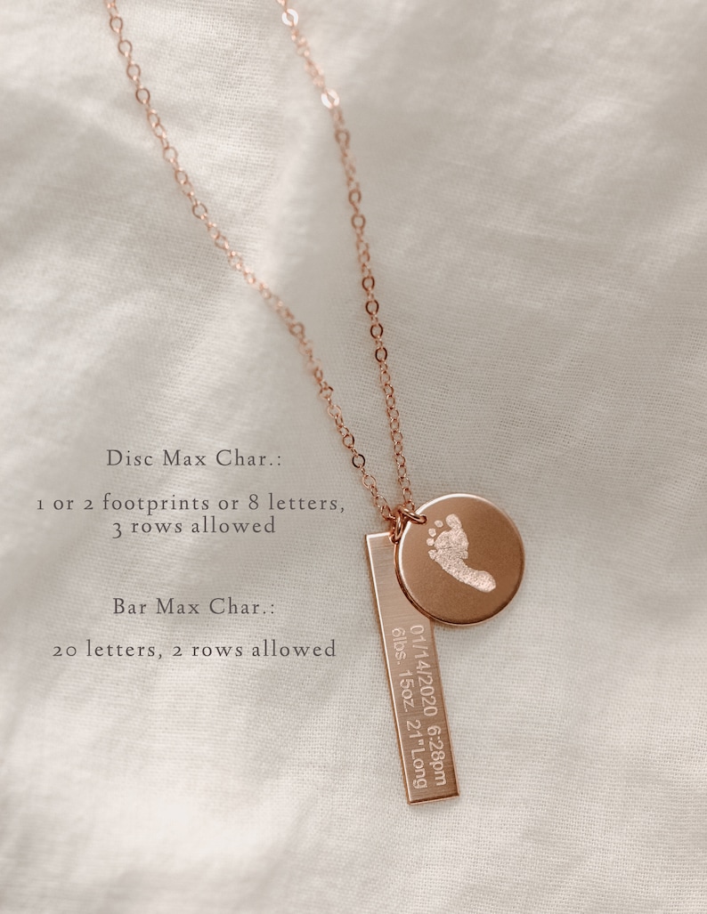 May include: A rose gold necklace with a small round disc charm and a rectangular bar charm. The disc charm has a footprint engraved on it. The bar charm has the text "01/14/2020 6:28pm 6lbs. 15oz. 21" Long" engraved on it. The necklace is on a white fabric background.