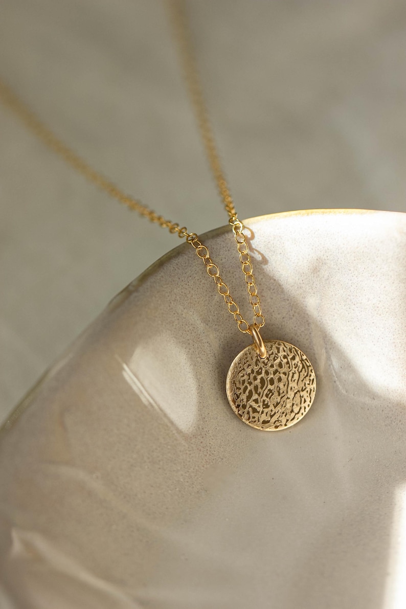 Pode incluir: Um colar dourado com um pendente circular texturizado. O pendente pende de uma corrente delicada. O colar &eacute; exibido sobre um fundo neutro, destacando o seu design e cor.