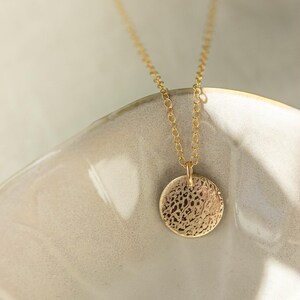 Pode incluir: Um colar dourado com um pendente circular texturizado. O pendente pende de uma corrente fina. A joia &eacute; apresentada numa superf&iacute;cie de cer&acirc;mica clara, oferecendo uma est&eacute;tica minimalista.