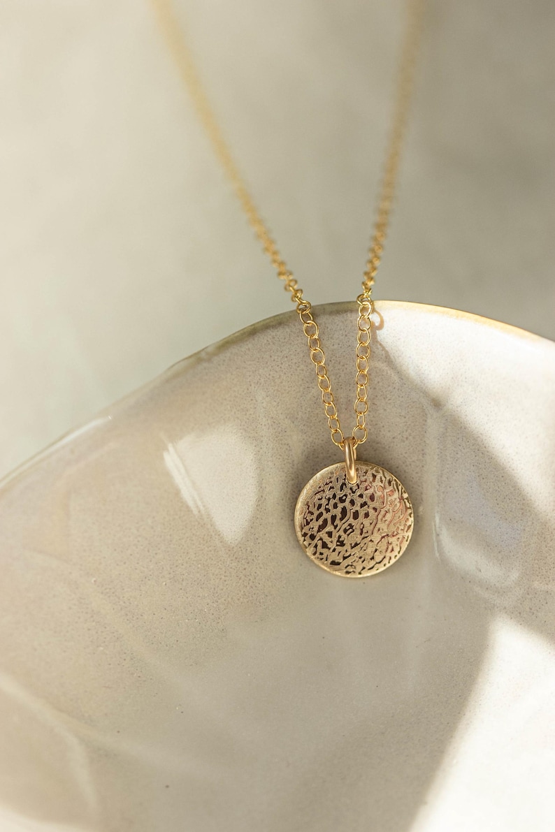 Pode incluir: Um colar dourado com um pendente circular texturizado. O pendente pende de uma corrente fina. A joia &eacute; apresentada numa superf&iacute;cie de cer&acirc;mica clara, oferecendo uma est&eacute;tica minimalista.