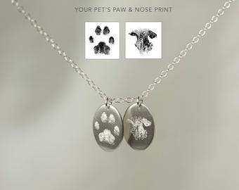 May include: A silver necklace with two oval pendants, each engraved with a pet's paw print and nose print. The text "YOUR PET'S PAW & NOSE PRINT" is above the pendants. The chain is delicate and the background is a soft gray.