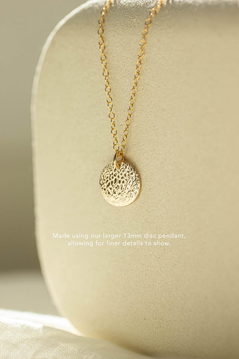 Pode incluir: Colar dourado com um pendente de disco texturizado de 13 mm. O pendente pende de uma corrente delicada. O texto na imagem diz: "Made using our larger 13mm disc pendant, allowing for finer details to show."