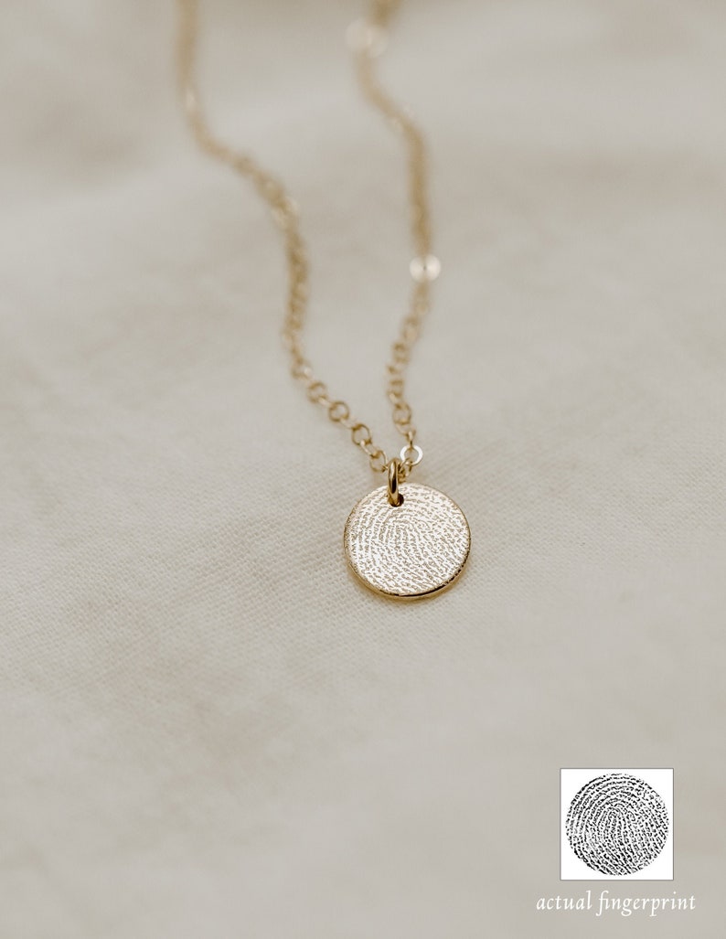 Puede incluir: Un collar de oro con un colgante redondo que presenta un dise&ntilde;o detallado de huella digital. El colgante tiene un di&aacute;metro de aproximadamente 2,5 cm. El collar est&aacute; sobre un fondo de tela blanca. La imagen tambi&eacute;n muestra un primer plano del dise&ntilde;o de la huella digital, etiquetado como "huella digital real".