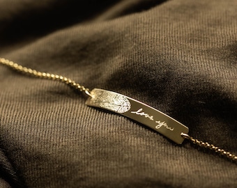 Puede incluir: Una pulsera dorada con un colgante rectangular. El colgante presenta un diseño de hoja y las palabras manuscritas "love you". La pulsera está sobre un fondo de tela marrón oscuro.
