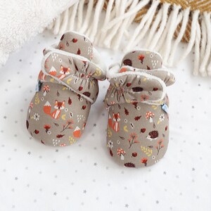 May include: A pair of baby booties with a woodland animal print. The booties are made of a soft, brown fabric with a white fleece lining. The print features foxes, hedgehogs, mushrooms, and leaves in shades of orange, red, yellow, and green.