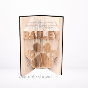 以下が含まれることがあります： ページを折りたたんで足跡のデザインと名前「BAILEY」を作成した本。 本は開いており、折りたたまれたデザインが明らかになっています。 テキスト「example shown」が一番下にあります。