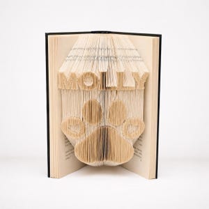 May include: A book with pages folded to create a three-dimensional sculpture. The top section features the name MOLLY in block letters, while the bottom section displays a large paw print design. The book has a black spine and cream-colored pages.