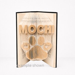 Peut inclure: Un livre dont les pages sont pliées pour former le mot "MOCHI" et une empreinte de patte. Le texte "example shown" est visible. Le livre a une tranche noire et les pages pliées sont beiges.