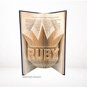 Op de afbeelding: Een gevouwen boekkunstsculptuur met de naam RUBY gevormd door nauwkeurig gevouwen pagina's. Het boek heeft een donkerblauwe kaft en crèmekleurige papieren pagina's die zijn bewerkt om een driedimensionale kroonvorm boven de tekst te creëren.