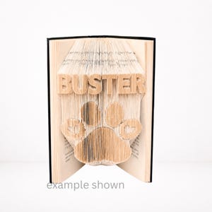 Peut inclure: Un livre ouvert dont les pages sont pliées pour révéler le nom "BUSTER" et une empreinte de patte. Le livre a une tranche noire et les mots "example shown" sont en bas. Le texte et l'empreinte de patte sont au centre.