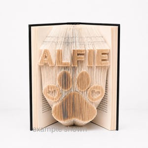 Könnte beinhalten: Eine Buchfaltkunst-Skulptur mit dem Namen ALFIE und einem Pfotenabdruck-Design, das durch präzises Falten der einzelnen Seiten eines Hardcover-Buches entsteht. Die cremefarbenen Papierseiten bilden die Buchstaben und die Pfotenform.