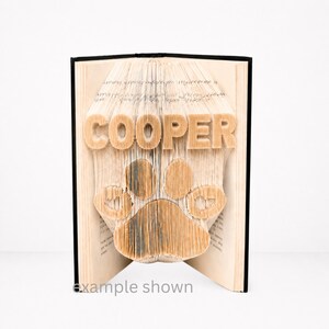 Puede incluir: Un libro abierto con una huella de pata y el nombre "COOPER" creado doblando las páginas. La huella de pata es el diseño central. El texto "example shown" está en la parte inferior.