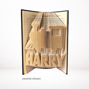 Op de afbeelding: Een boeksculptuur met een treinontwerp en de naam "HARRY" uitgesneden in de pagina's. Het open boek toont het beeld dat in de pagina's is gesneden. De tekst "examle shown" staat onderaan.