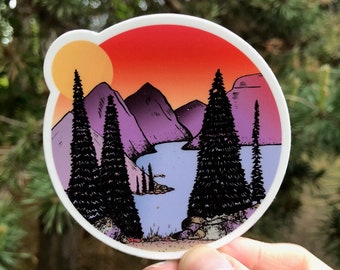 May include: A round sticker with a sunset scene featuring a lake, mountains, and pine trees. The sky is a gradient of orange, pink, and purple. The sun is a bright yellow circle in the upper left corner.