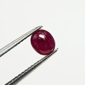 May include: A deep red oval-shaped gemstone held by tweezers. The gemstone is smooth and polished.