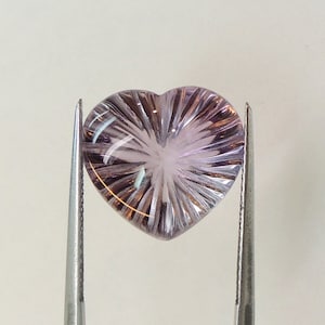 May include: A heart-shaped amethyst gemstone with a carved, starburst pattern. The gemstone is a pale purple colour and is held in place by two metal tweezers.