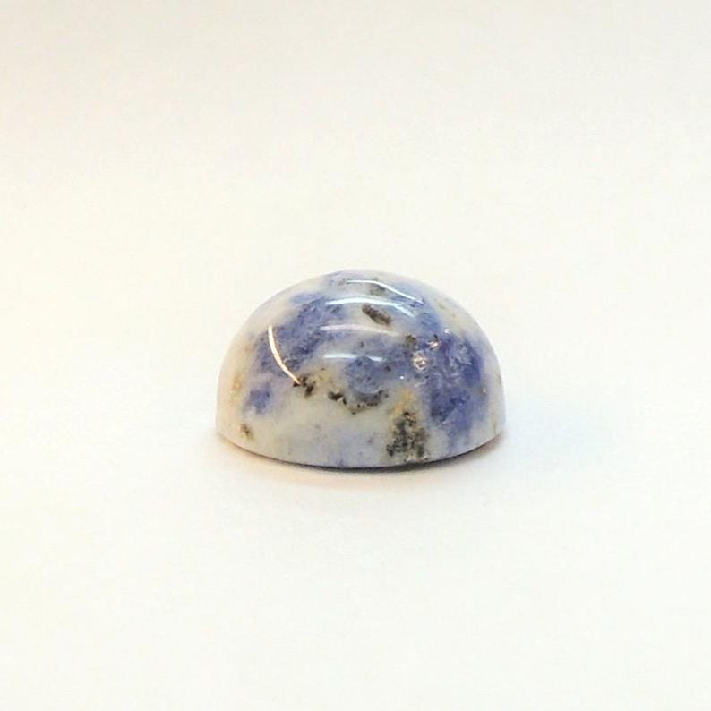 Op de afbeelding: Een gepolijste, cabochon-geslepen edelsteen met een afgeronde koepelvorm. De steen heeft een mix van witte, blauwe en grijze kleuren, met een gespikkeld patroon. De edelsteen is geplaatst tegen een effen witte achtergrond.