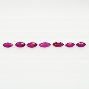May include: Seven marquise-shaped ruby gemstones, each with a deep red hue, are arranged in a row on a white background.