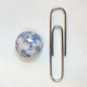 Op de afbeelding: Een ronde, gepolijste steen met een blauw en wit gevlekt patroon, naast een grote zilveren paperclip. De steen heeft een glanzende afwerking. De paperclip is gemaakt van glanzend metaal.