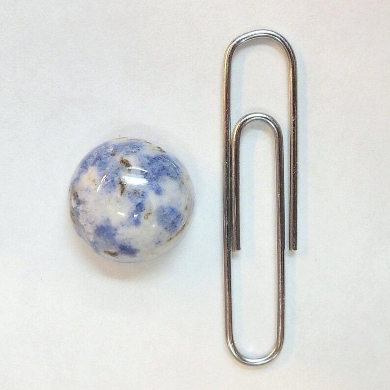 Op de afbeelding: Een ronde, gepolijste steen met een blauw en wit gevlekt patroon, naast een grote zilveren paperclip. De steen heeft een glanzende afwerking. De paperclip is gemaakt van glanzend metaal.