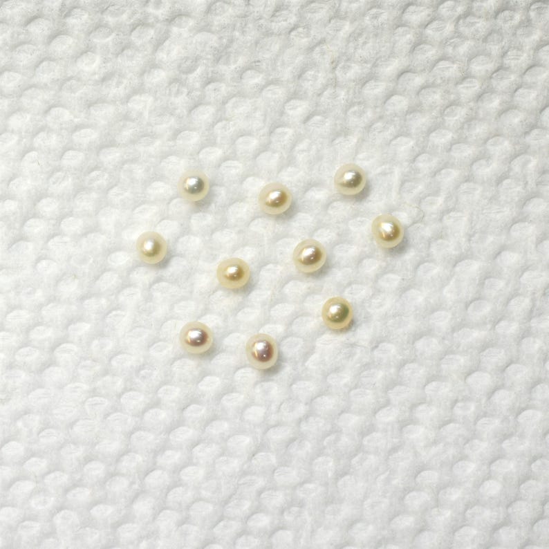 May include: A collection of ten white pearls of various sizes, arranged in a heart shape on a white textured surface.