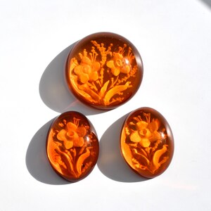 May include: Three amber glass cabochons with floral designs. The cabochons are oval and round in shape. The flowers are carved into the glass and are a light orange color.