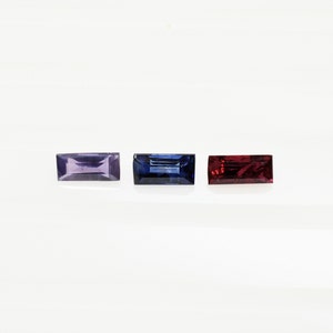 May include: Three rectangular gemstones, one purple, one blue, and one red, are displayed on a white surface.