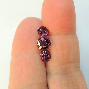 May include: Three round, faceted, purple gemstones on a finger. The gemstones are arranged vertically, with the largest at the bottom.