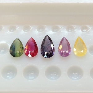 May include: Five pear-shaped gemstones in a variety of colours, including green, red, purple, pink, and yellow, are displayed in a white tray with individual compartments.