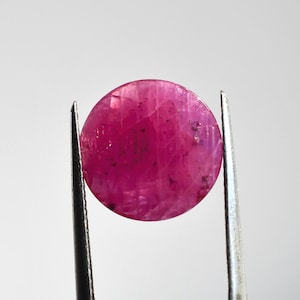 Peut inclure: Une pierre précieuse de rubis ronde et polie, tenue par une pince. Le rubis est d'un rouge-rosé profond avec des inclusions et des stries internes visibles. La pierre est posée sur un fond blanc uni.