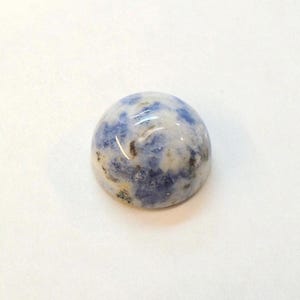 Op de afbeelding: Een gepolijste, ronde edelsteen met een mix van blauwe, witte en bruine kleuren. De steen heeft een glad, gewelfd oppervlak en lijkt een cabochon te zijn, geschikt voor het maken van sieraden of verzamelen. De steen staat tegen een witte achtergrond.