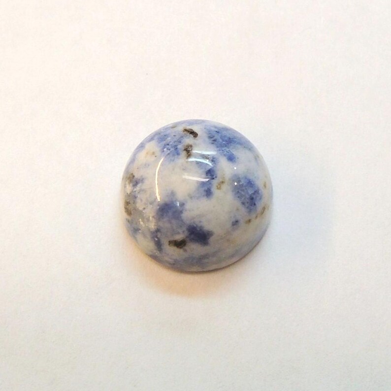 Op de afbeelding: Een ronde, gepolijste edelsteen met een mix van witte en blauwe kleuren. De steen heeft een glad, gewelfd oppervlak met een gespikkeld patroon. Kleine donkere vlekken zijn verspreid.