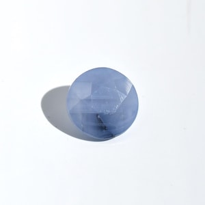 May include: A round, pale blue gemstone with multiple facets. The polished surface of the gem reflects light, creating a shimmering effect. The stone is set against a white background, with a soft shadow to the left.