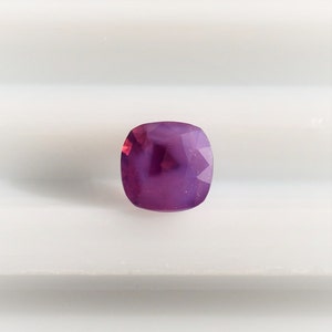 May include: A cushion-cut gemstone with a purple hue and a slight pink tint. The gemstone is faceted and has a high polish.