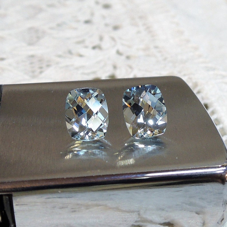 Puede incluir: Dos piedras preciosas de aguamarina azul claro, cada una con un corte de coj&iacute;n, se muestran sobre una superficie plateada.