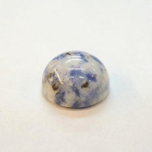 Op de afbeelding: Een gepolijste, koepelvormige edelsteen met een gemarmerd uiterlijk. De steen heeft een mix van witte, blauwe en bruine tinten, waardoor een uniek patroon ontstaat. Het oppervlak is glad en reflecterend, wat wijst op een hoog niveau van vakmanschap. Deze edelsteen kan worden gebruikt in sieraden of als decoratief item.