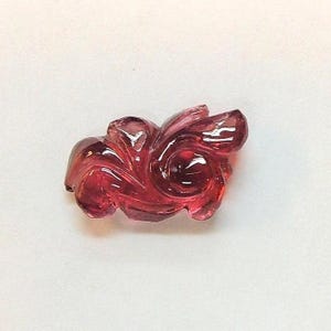 May include: A carved, translucent, deep red gemstone with a swirling, floral design. The polished surface reflects light, highlighting the intricate details of the carving. The gemstone is set against a white background.