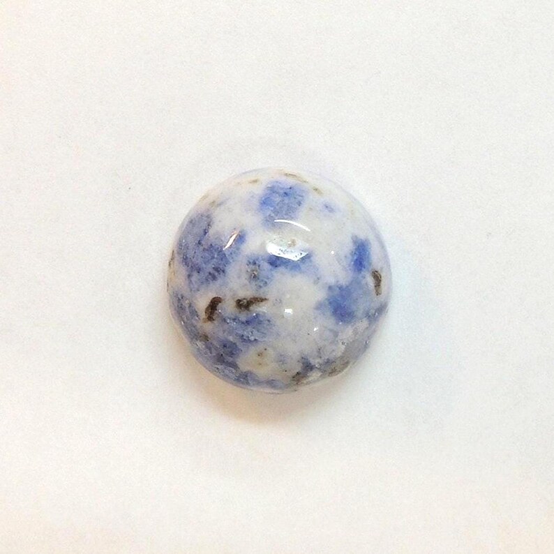 Op de afbeelding: Een ronde, gepolijste edelsteen met een gespikkeld patroon van blauw en wit. De steen heeft een glad, glanzend oppervlak en een mix van licht- en donkerblauwe vlekken. De edelsteen staat tegen een witte achtergrond.