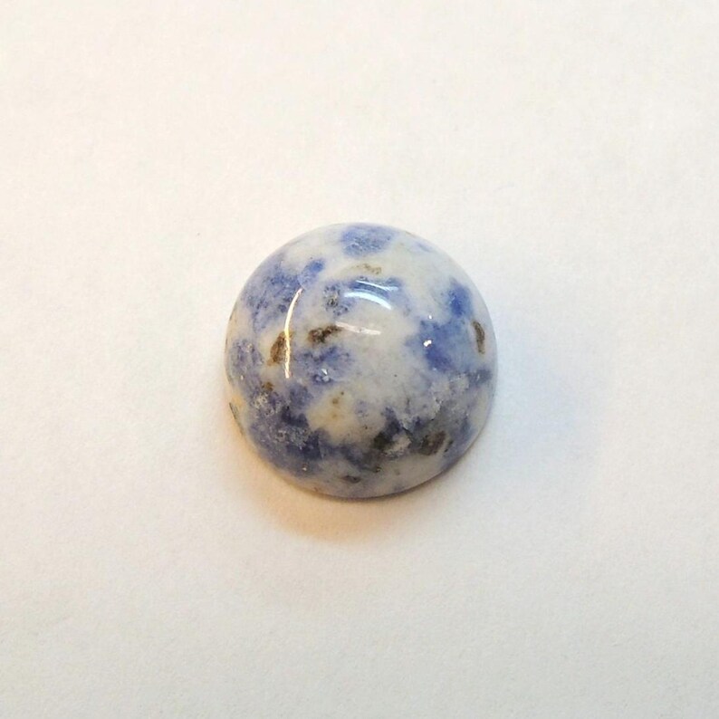 Op de afbeelding: Een ronde, gepolijste edelsteen met een mix van blauwe, witte en bruine kleuren. De steen heeft een glad, glanzend oppervlak en een gespikkeld patroon. De edelsteen is geplaatst tegen een witte achtergrond.